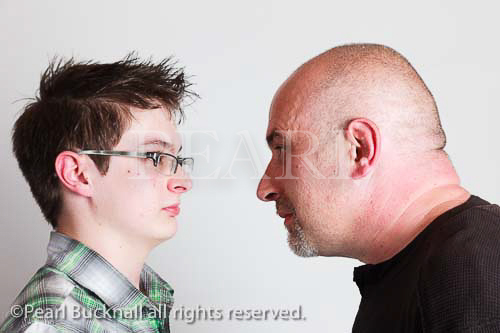 Head and shoulders portrait of a father and teenage 
son facing each other and dad looking cross