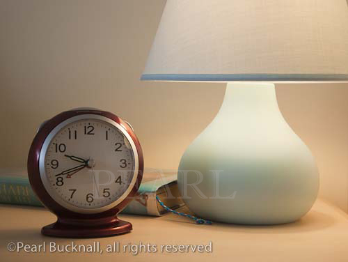 Close up of an alarm clock with lit bedside table lamp 
and book in the evening

Keywords: light bedroom nobody closeup