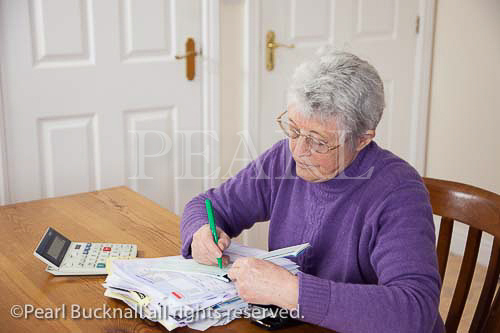 Senior woman pensioner with a big pile of bills on the 
table writing a cheque to pay. UK Britain MR 10/06