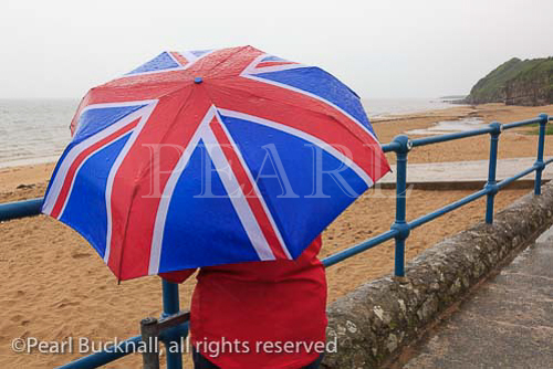 A lone woman looks out to sea on the seafront of 
deserted seaside resort with a Union Jack umbrella 
during very wet British summer weather. Benllech, 
Anglesey, North Wales, UK. 