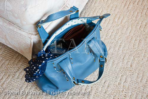 Women's blue leather handbag with zip open showing 
contents on the floor carpet beside a chair