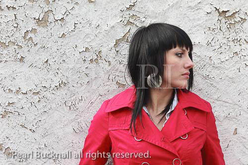 Young woman wearing a red coat standing by a white 
wall with peeling paint. UK Britain Europe. 