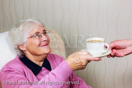 Senior woman looking up and smiling at a carer giving 
her a cup of tea. England, UK, Britain. MR 13/29, MR 
13/30