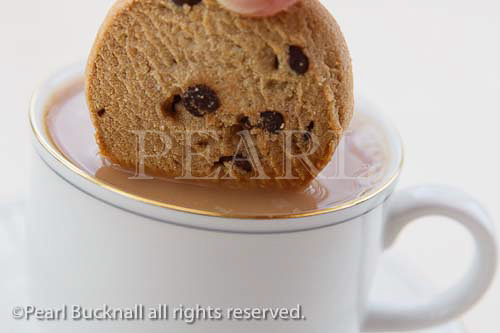 Person dunking a chocolate chip biscuit into a cup of 
tea in a white china teacup. England, United Kingdom, 
Britain