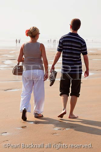 Young couple walking on sandy beach, man 
barefoot with trousers rolled up carrying shoes. 
Bude, Cornwall, England, UK, Britain, Europe. 