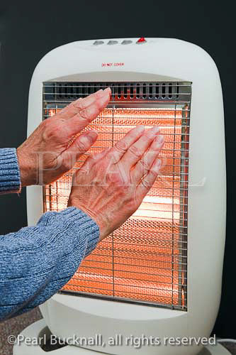 Elderly senior woman warming hands in front of a 
low energy Halogen room heater. England UK 
Britain