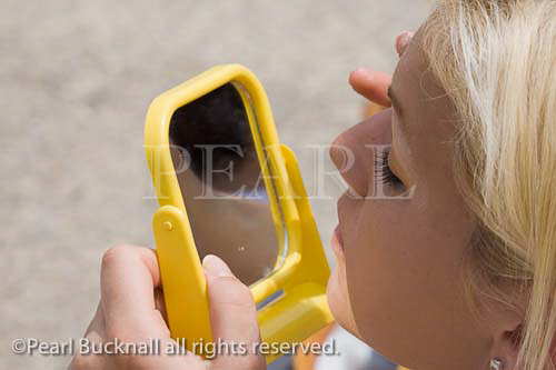 Attractive young blonde haired woman using a 
handheld mirror applying eye face make up