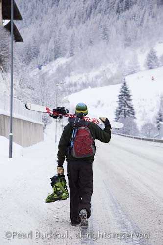 Skier carrying skis and ski boots walking down snow 
covered Alpine road in mid winter. St Anton am 
Arleberg, Tyrol, Austria, Europe