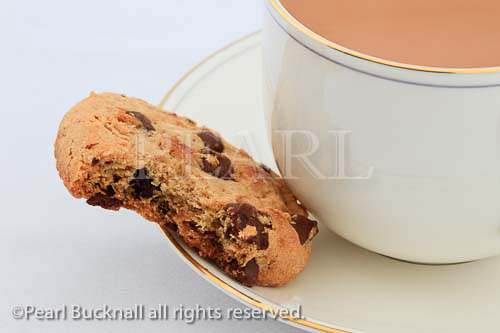 Cup of tea and a chocolate chip biscuit with a bit taken 
out on the saucer. Studio still life. 
