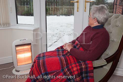Senior woman with a rug on her knees keeping
warm indoors by an electric fire with snow outside
the window in winter. UK Britain Europe