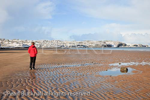 Person wearing a red jacket walking on an empty 
sandy beach at low tide in winter. Benllech, Isle of 
Anglesey, North Wales, UK, Europe. 

Keywords: coast scene snowfall Britain British weather 
Welsh Ynys Mon