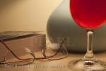 Glass of red wine with lit bedside table lamp 
paperback book and spectacles in the evening

Keywords: light still life home homely concept