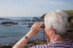 Senior woman looking out to sea through binoculars 
on the rugged south coast at the most southerly point 
of Britain. Lizard Point, Cornwall, England, UK