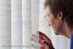Senior woman being nosy peeping through window 
blinds to look outside. England, United Kingdom, 
Britain